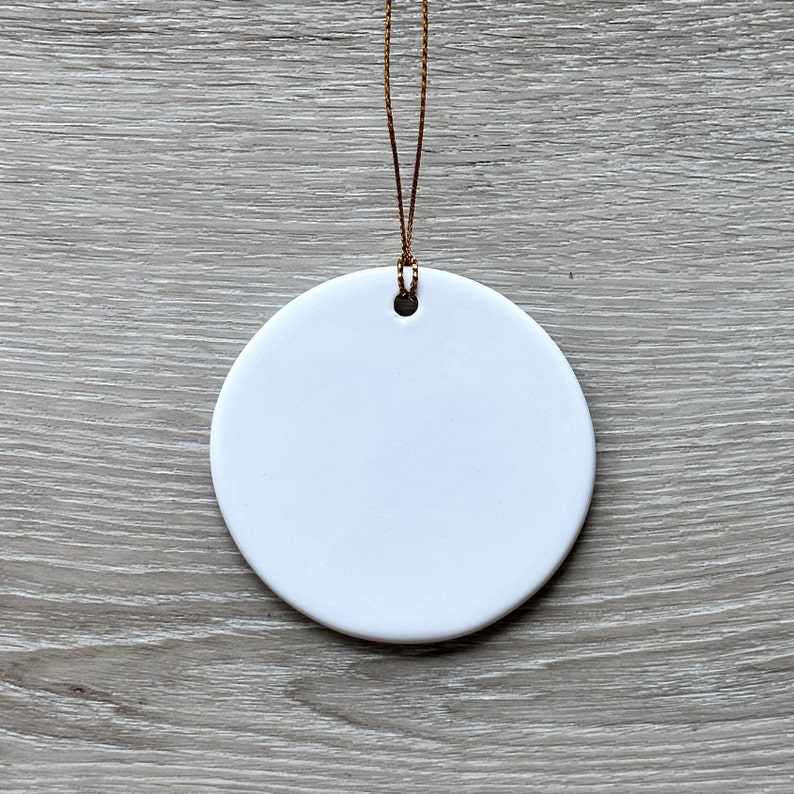 May include: A white ceramic ornament with a gold string attached. The ornament is round and has a hole in the top for hanging.