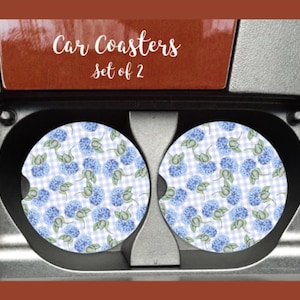 May include: Two car coasters with a blue hydrangea floral pattern on a white and light blue gingham background. The coasters are set in a car's cup holders. The text "Car Coasters set of 2" is visible.
