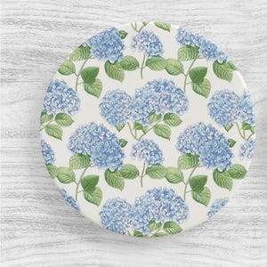 May include: A white ceramic plate with a blue and green floral pattern. The plate features a repeating pattern of blue hydrangea flowers with green leaves.