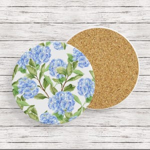 May include: Two round ceramic coasters with a cork backing. One coaster features a white background with a blue watercolor floral pattern of hydrangeas. The other coaster is plain cork.