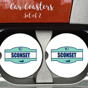 May include: Two white car coasters with a blue and green design. The design features the text "Sconset" and a small image of an island. The coasters are in a car cup holder.
