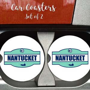 May include: Set of two white car coasters with a blue and green design featuring the text "NANTUCKET" and a small island graphic. The coasters are in a car cup holder.