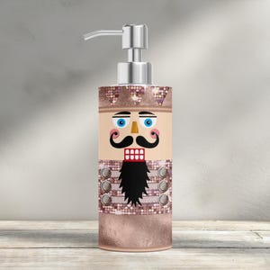May include: A decorative soap dispenser featuring a nutcracker design. The dispenser has a silver pump and a cylindrical body with a festive illustration of a nutcracker soldier. The design includes a black mustache, a black beard, and blue eyes.