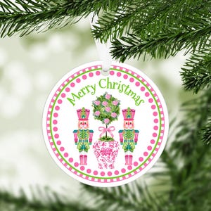 May include: White round Christmas ornament with pink and green polka dots. The ornament features two nutcracker figures flanking a pink and green topiary with the text "Merry Christmas" in green.