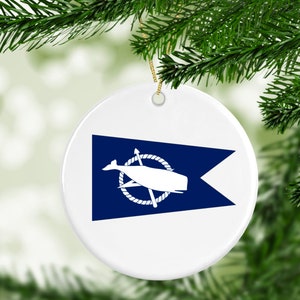 May include: A white ceramic Christmas ornament with a blue and white nautical flag design. The flag features a white whale with a compass rose and a white anchor.