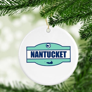 May include: White ceramic Christmas ornament with a blue and green design. The ornament features the text "NANTUCKET" and a small outline of an island.