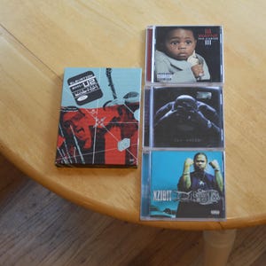 May include: A collection of music CDs on a wooden table. The CDs feature album art with images of artists and text. One CD case has a baby on the cover. Another CD case has a person in a dark room. The last CD case has a person in a blue background.