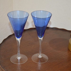 May include: Two elegant blue-tinted stemmed glasses with a clear base and stem. The glasses have a conical bowl shape. They are placed on a dark brown, round table. A gold-colored lamp base is visible in the background.