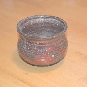 May include: A handmade ceramic bowl with a rustic appearance. The bowl features a rounded shape with a wide opening and a textured surface. The colors include shades of brown, gray, and reddish-orange, with white speckles.