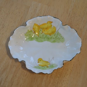 May include: A white ceramic dish with a scalloped edge and a black trim. The dish features a hand-painted design of three yellow chicks in a grassy field.