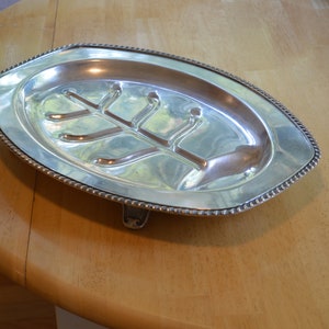 May include: A silver serving platter with a tree-shaped design and a beaded edge. The platter has three feet and is oval shaped.
