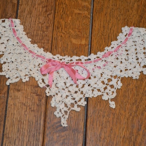 May include: A white crocheted lace collar with a pink satin ribbon tied in a bow at the center.