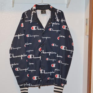 May include: A navy blue jacket with a white and red "Champion" logo pattern. The jacket has a button-up front and a zippered pocket on the left side.