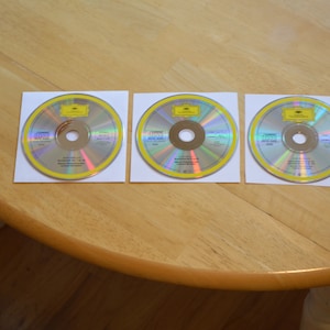 May include: Three silver compact discs with yellow borders and a yellow label. The label has the text "Deutsche Grammophon" and "The Complete Symphonies" in black text.