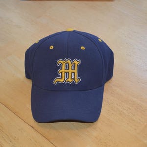 May include: A navy blue baseball cap featuring an embroidered letter M in yellow with a white outline on the front panel. The cap includes yellow eyelets on the crown and a yellow button at the top center, constructed with a structured six-panel design.