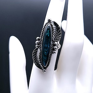May include: A silver-toned ring with an oval, iridescent, teal-colored stone. The ring features a decorative leaf and bead design around the stone. The ring is displayed on a white surface.