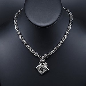 May include: A silver chain necklace with a square pendant featuring a detailed design. The chain is a woven style.