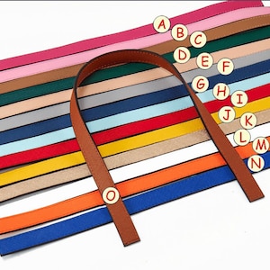Puede incluir: Un conjunto de 15 correas de cuero de colores con un asa de cuero marr&oacute;n. Las correas est&aacute;n dispuestas en un patr&oacute;n de arco&iacute;ris y est&aacute;n etiquetadas con las letras de la A a la O.