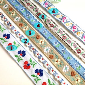 Könnte beinhalten: Sechs verschiedene Arten von floral besticktem Besatzband in verschiedenen Farben, darunter Blau, Weiß, Braun und Rosa. Der Besatz ist etwa 1 Zoll breit und zeigt eine Vielzahl von Blumendesigns.