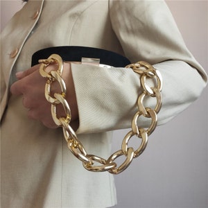 May include: A gold chain handbag strap with large, chunky links. The strap is attached to a black clutch bag.