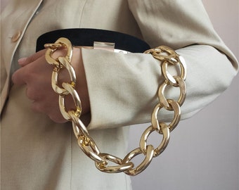 May include: A gold chain handbag strap with large, chunky links. The strap is attached to a black clutch bag.