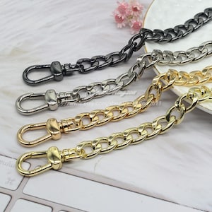 Puede incluir: Primer plano de cuatro correas de cadena metálicas en negro, plateado y dorado. Cada cadena tiene un cierre de mosquetón en un extremo. Las cadenas son gruesas y tienen un diseño de eslabones de bordillo. Las cadenas doradas tienen un acabado brillante.