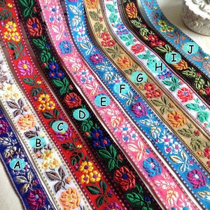 Op de afbeelding: Tien kleurrijke geborduurde lintjes met bloemenpatronen. De lintjes zijn in een rij van links naar rechts gerangschikt, met kleuren die variëren van donkerblauw tot roze. Elk lintje heeft een ander bloemenpatroon, en de kleuren zijn levendig en opvallend.