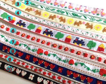 May include: Ten different embroidered ribbon trims with various designs, including flamingos, dogs, houses, trains, strawberries, and birds. The ribbons are white with colorful embroidery.