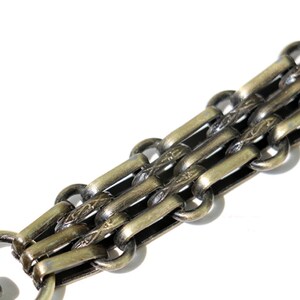 May include: A close-up of a gold-toned chain with a decorative pattern. The chain is made of multiple interwoven links.