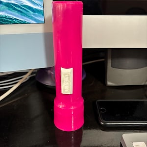 May include: A bright pink, cylindrical flashlight with a silver rectangular label that reads "FUR READY". The flashlight has a wider base and a narrower top. The flashlight is standing upright.