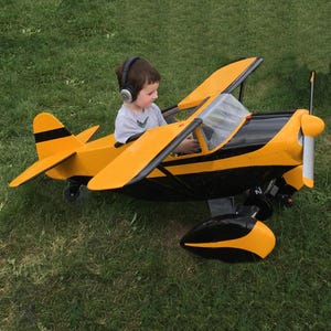 Könnte beinhalten: Ein gelb-schwarzes Spielzeugflugzeug mit Propeller und Flügeln. Das Spielzeugflugzeug hat ein schwarzes Streifendesign und eine klare Windschutzscheibe. Ein Kind mit Kopfhörern sitzt im Cockpit des Flugzeugs auf einer Wiese.