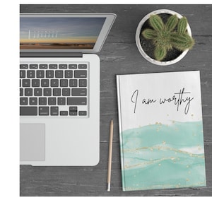 May include: A white journal with a teal and gold watercolor design and the text "I am worthy" written in black cursive.