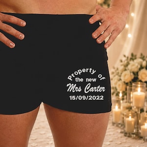 May include: Black boxer shorts with white text reading "Property of the new Mrs Carter 15/09/2022". The shorts are a solid black colour and likely made of a soft material. The text is in a script font.