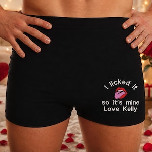 Może przedstawiać: Czarne bokserki z grafiką czerwonych ust z różowym językiem. Napis brzmi: I licked it so it's mine Love Kelly. Wzór znajduje się w dolnej prawej części odzieży, która posiada szeroki, jednolity czarny pas w talii.