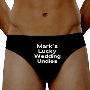 以下が含まれることがあります： 白い文字で「Mark's Lucky Wedding Undies」と書かれた黒い下着。