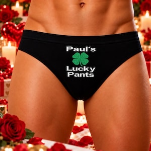 May include: Black underwear featuring a green four-leaf clover graphic centred between the white text Paul's and Lucky Pants. The garment has a solid black waistband and a classic brief cut, displaying the printed design clearly on the front.