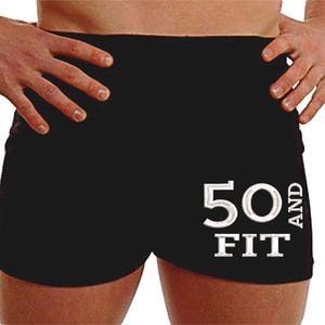 May include: Black athletic shorts with the text "50 AND FIT" embroidered in white. The shorts are form-fitting and appear to be made of a stretchy material. The image shows the shorts from the waist down.