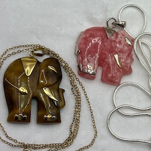 Puede incluir: Dos colgantes de elefante en cadenas. Un colgante está hecho de piedra de ojo de tigre marrón y el otro de piedra rosa.