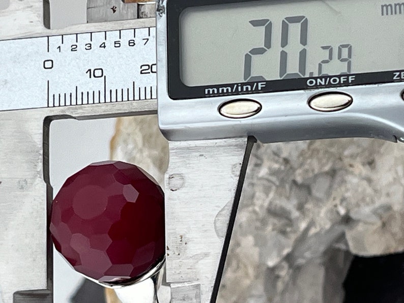 Op de afbeelding: Een close-up van een diep rode gefacetteerde edelsteen die wordt gemeten met een digitale schuifmaat. De schuifmaat geeft 20,29 mm aan.