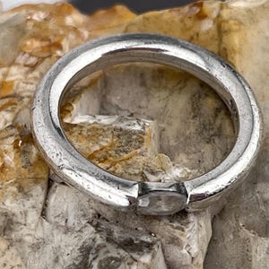 May include: A silver ring with a rectangular, clear stone set in the band. The ring is round and has a textured surface. The ring is set against a light brown and tan stone background.