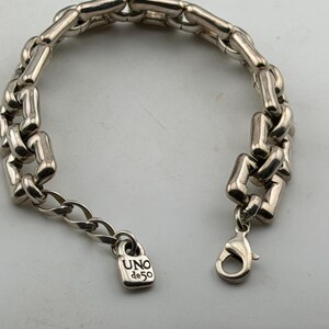 May include: Silver bracelet with a chunky chain link design. The bracelet has a lobster clasp and a small padlock charm engraved with "UNO de 50". A fashionable accessory.
