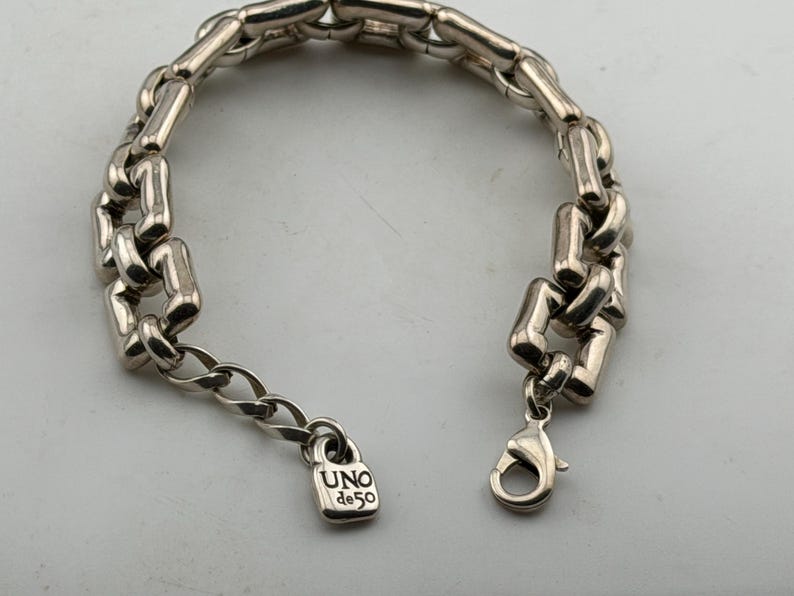 May include: Silver bracelet with a chunky chain link design. The bracelet has a lobster clasp and a small padlock charm engraved with "UNO de 50". A fashionable accessory.