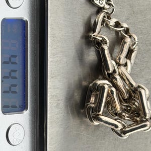 May include: A silver chain bracelet with a rectangular link design, resting on a digital scale. The bracelet has a small tag with the text "UNO de 50". The scale displays a weight of 4.44.
