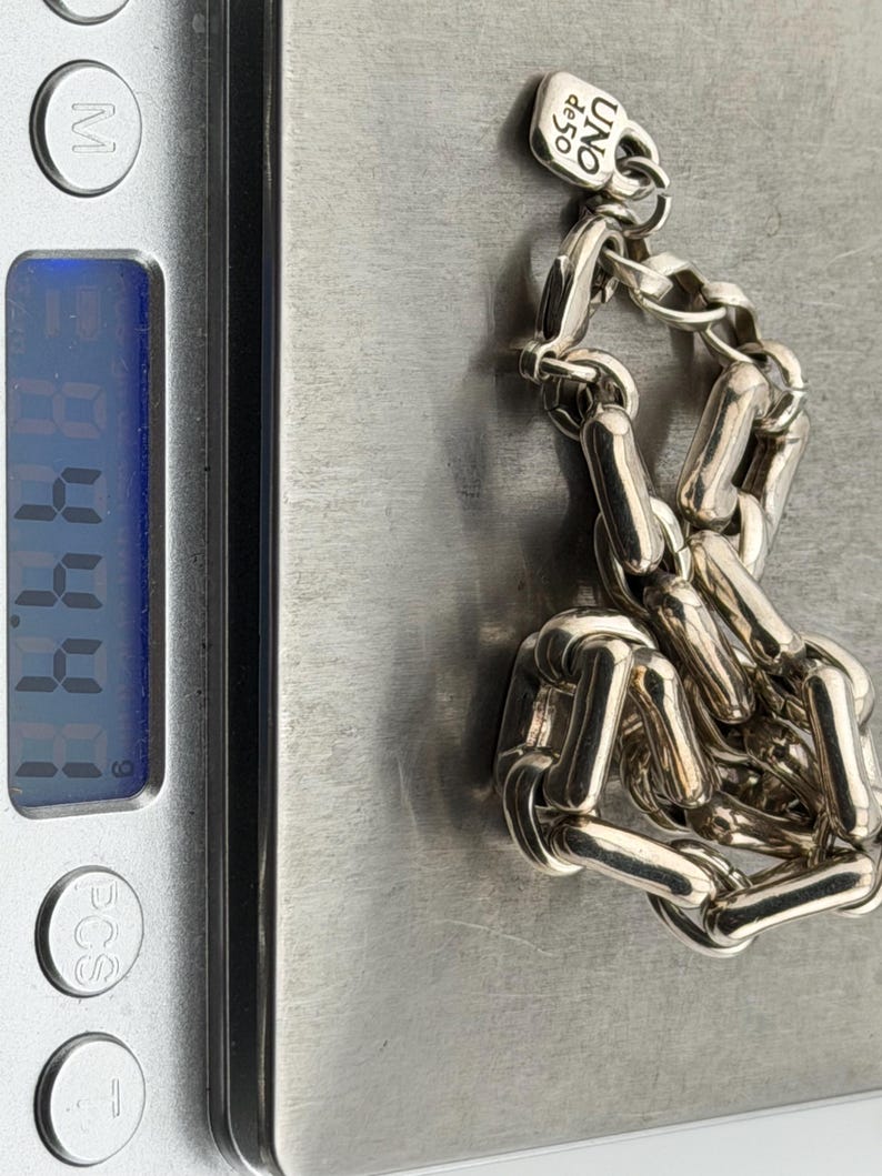 May include: A silver chain bracelet with a rectangular link design, resting on a digital scale. The bracelet has a small tag with the text "UNO de 50". The scale displays a weight of 4.44.