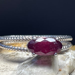 May include: A silver ring featuring a deep red oval gemstone set in the centre. The ring band is twisted and has a delicate design.