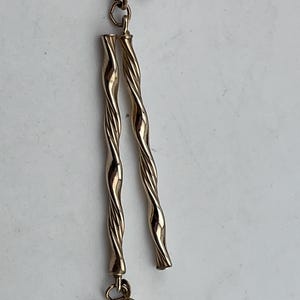 Könnte beinhalten: Goldfarbene Hängeohrringe. Jeder Ohrring hat einen Stecker, eine kleine Verbindungsschlaufe und ein Paar verdrehter, länglicher Stäbe. Die Ohrringe haben eine polierte Oberfläche und ein schlichtes, elegantes Design.