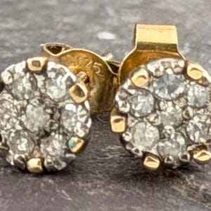 May include: A pair of gold stud earrings, each featuring a circular cluster of seven small, clear, round-cut stones set in a gold frame. The back of the earring post is clearly stamped with the number 925, indicating the metal composition.