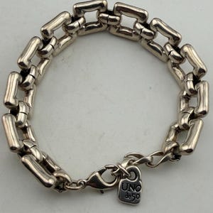 May include: Silver bracelet with a series of rectangular links. The bracelet has a lobster clasp and a small tag with the text "UNO de 50". The links are connected to form a chain.