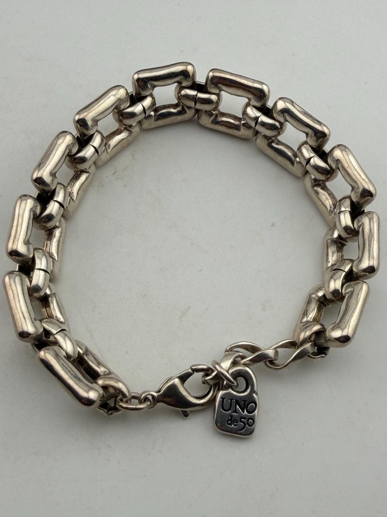 May include: Silver bracelet with a series of rectangular links. The bracelet has a lobster clasp and a small tag with the text "UNO de 50". The links are connected to form a chain.
