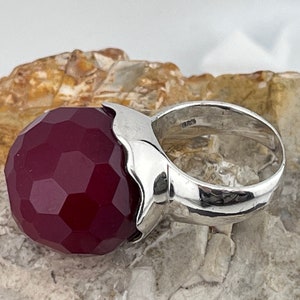 Op de afbeelding: Een zilveren ring met een grote, gefacetteerde, donkerrode edelsteen. De ring is in een eenvoudige, moderne stijl gezet.
